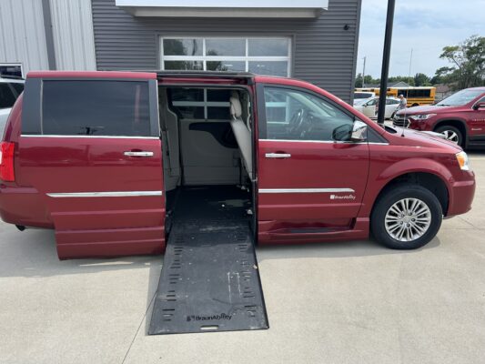 Red minivan with a deployed wheelchair ramp extending from the side door, BraunAbility branding visible on the ramp, parked outside a building with other vehicles nearby.