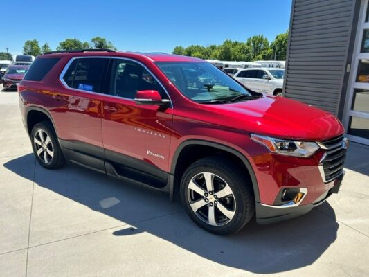 Red Chevrolet Traverse SUV parked on a dealership lot beside a gray building with other vehicles in the background on a sunny day.