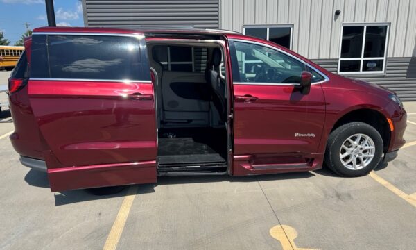 Maroon BraunAbility wheelchair-accessible minivan with side-entry ramp deployed in a parking lot, sliding door open, interior visible beneath roof line.