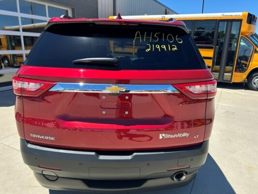 Rear view of a red Chevrolet Traverse LT with BraunAbility branding in a parking lot; rear window shows writing and a chrome bumper, with a yellow bus in the background.