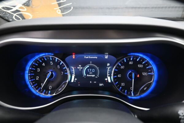 Blue-lit car dashboard: left tachometer, right speedometer, center digital display shows Fuel Economy and 17.0 MPG average, raindrop-speckled windshield visible above.