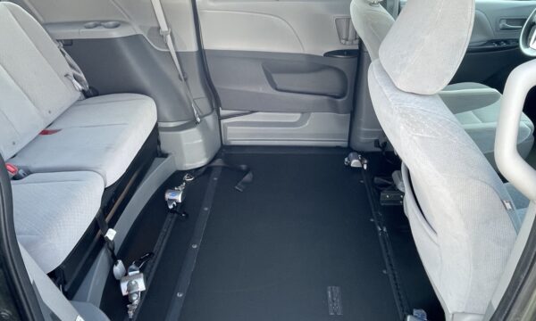 IMG_0391 Interior of a light-gray passenger van with the second-row seat removed, exposing a flat black floor and wheelchair tie-down anchors along the sides for securement.