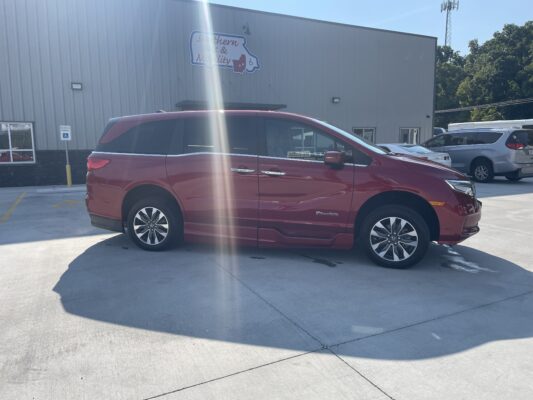 Red BraunAbility minivan parked in a lot in front of a metal building, near a handicap parking sign and other vehicles.