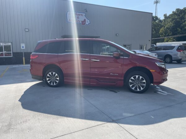 IMG_0438 Red BraunAbility minivan parked in a lot in front of a metal building, near a handicap parking sign and other vehicles.