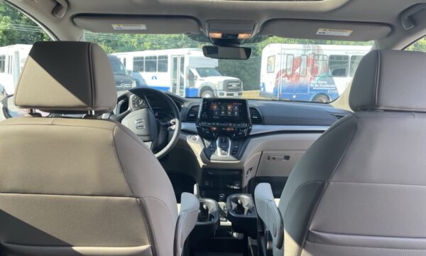 IMG_0446 Interior view from the back seat of a car, showing the dashboard with a touchscreen, steering wheel, gear shifter, and two front seats; buses are visible outside through the windshield.