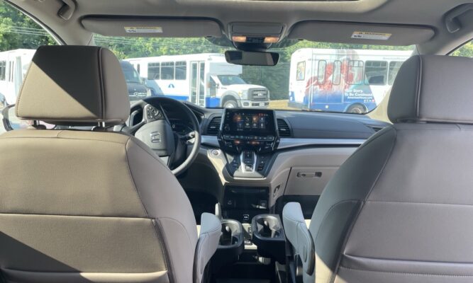 Interior view from the back seat of a car, showing the dashboard with a touchscreen, steering wheel, gear shifter, and two front seats; buses are visible outside through the windshield.