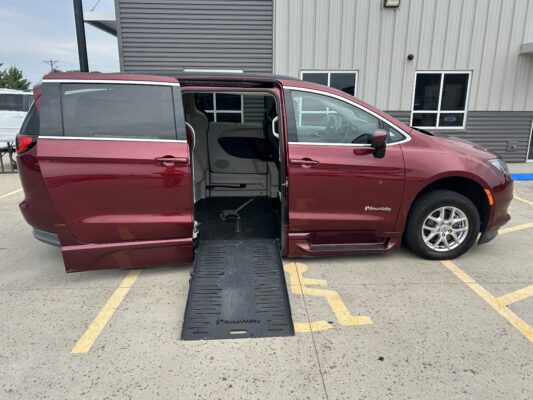Maroon BraunAbility wheelchair-accessible van with ramp extended and side door open in a parking lot by a building district office complex.