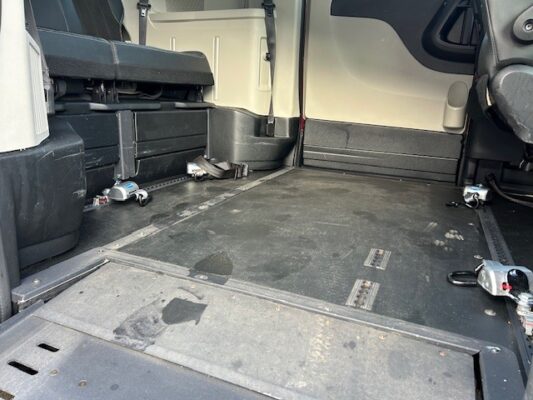 Cargo van interior with a bare floor, folded passenger seat on the left, wheel wells, and metal tie-downs along the floor.