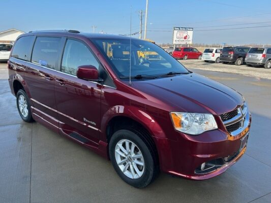 Maroon Dodge Grand Caravan minivan parked on a dealership lot with other cars in the background.