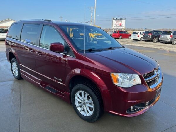 Passenger side angle Maroon Dodge Grand Caravan minivan parked on a dealership lot with other cars in the background.