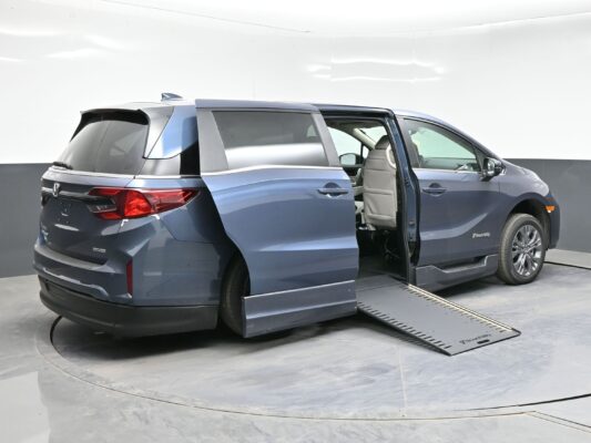 Blue minivan with both side and rear doors open, showing a lowered ramp extending from the side for wheelchair access in a clinical testing space.