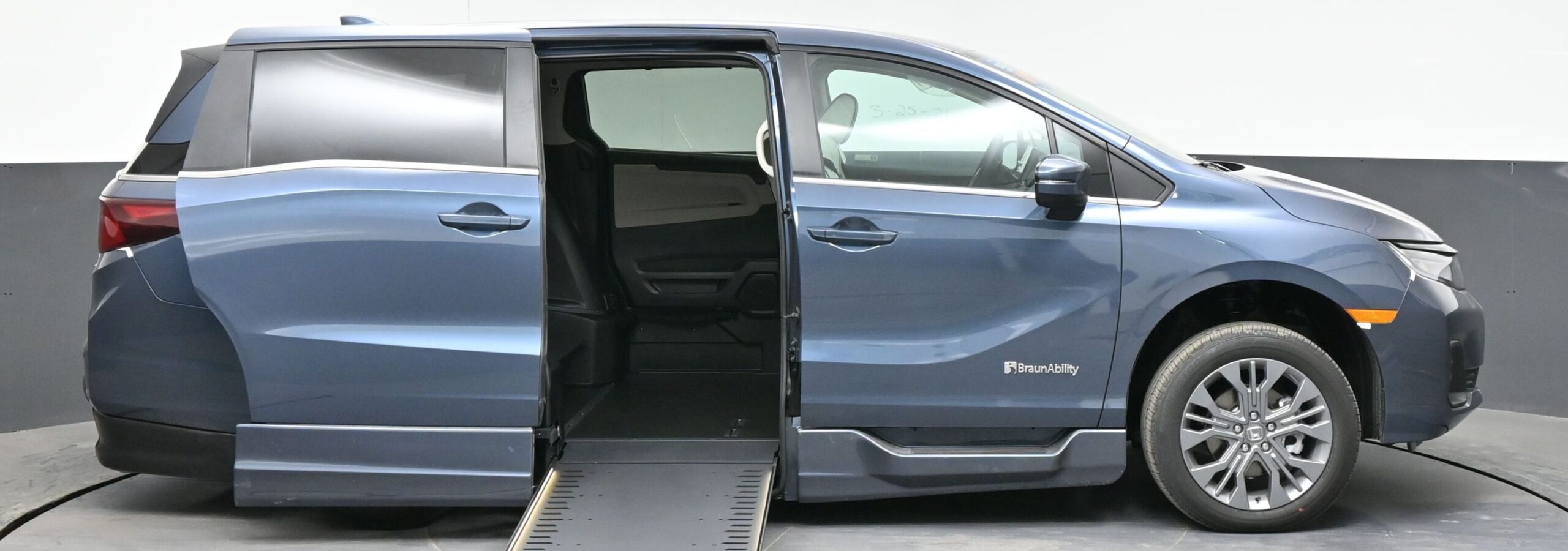Blue minivan with a side-entry ramp extended to the floor, BraunAbility logo visible in a studio setting.