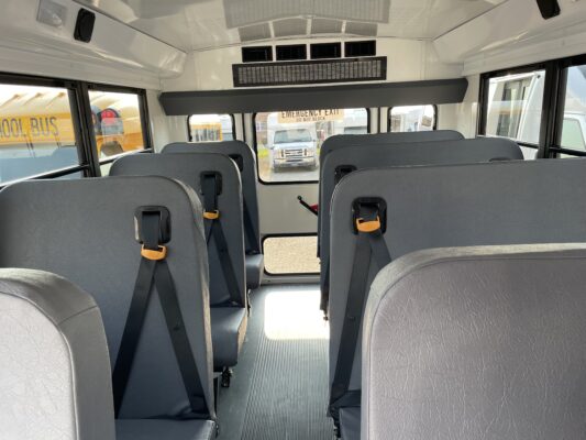 Aisle view inside a school bus with gray seats and orange buckle latches, emergency exit sign visible through the windows.