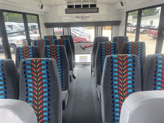 Interior of a school bus with bright patterned seats, a narrow aisle, and large windows showing parked cars outside.