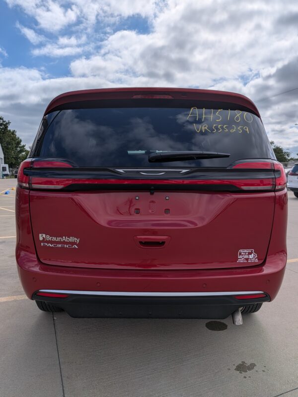 Red minivan rear view with BraunAbility Pacifica branding on left bumper and a window sticker on the right window against a cloudy sky backdrop
