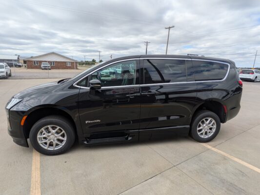 Side view of a black BraunAbility SUV parked in a lot, with a brick building and chain-link fence in the background under a cloudy sky.