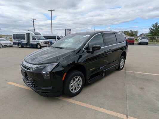 Black minivan parked diagonally in a lot with white vans and a cloudy sky in the background, foreground focus on the vehicle's front end and wheel.