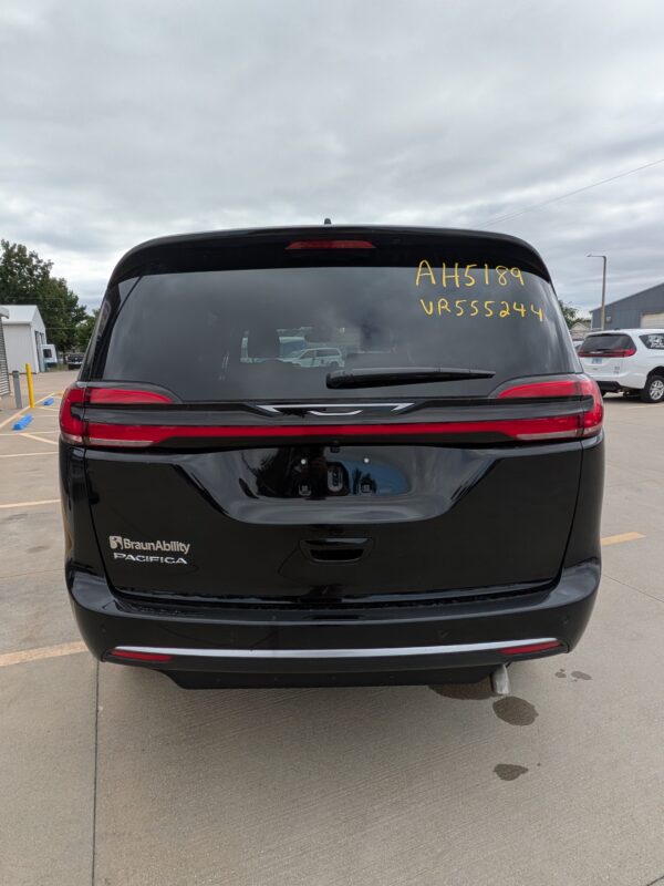 PXL_20260922_201345664 Rear view of a black BraunAbility Pacifica minivan in a parking lot, with yellow writing on the rear window (AH5189 VR55524). The vehicle has red tail lights and a rear wiper.