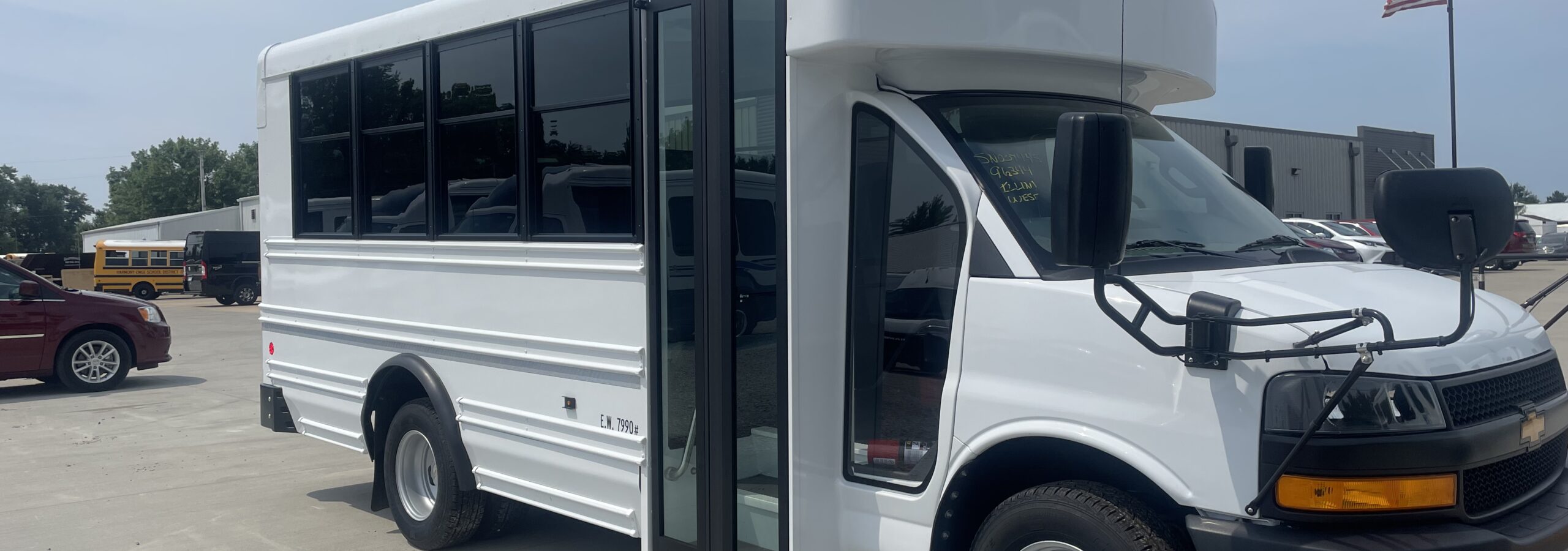 White school-bus shaped vehicle mounted on a truck chassis, labeled TRANS TECH, parked in a lot with an American flag in the background