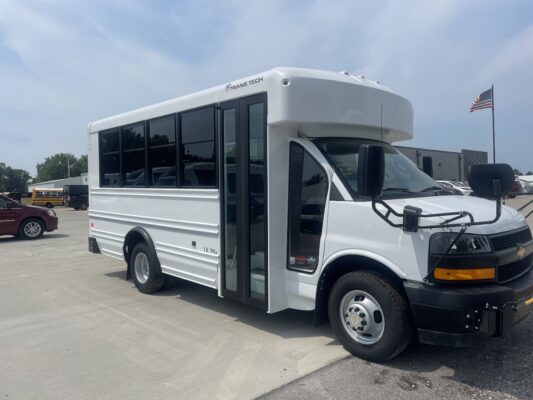 White school-bus shaped vehicle mounted on a truck chassis, labeled TRANS TECH, parked in a lot with an American flag in the background