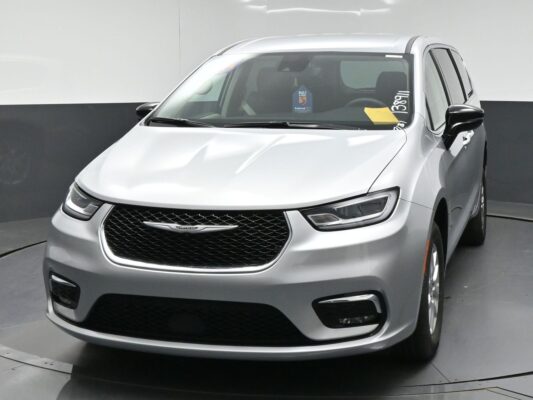 Front three-quarter view of a silver Chrysler minivan in a showroom, showing the glossy grille and headlights.