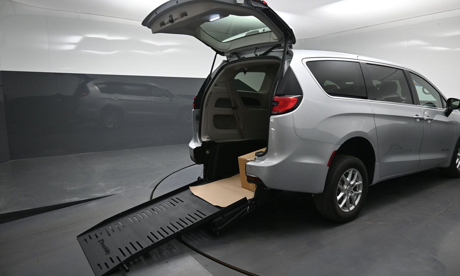 Silver minivan with rear hatch open, ramp extended to the floor, and a box inside the cargo area on a service lift.