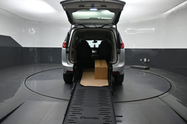 Back view of a silver minivan with its rear wheelchair ramp deployed, a cardboard box on the ramp in a white studio setting.
