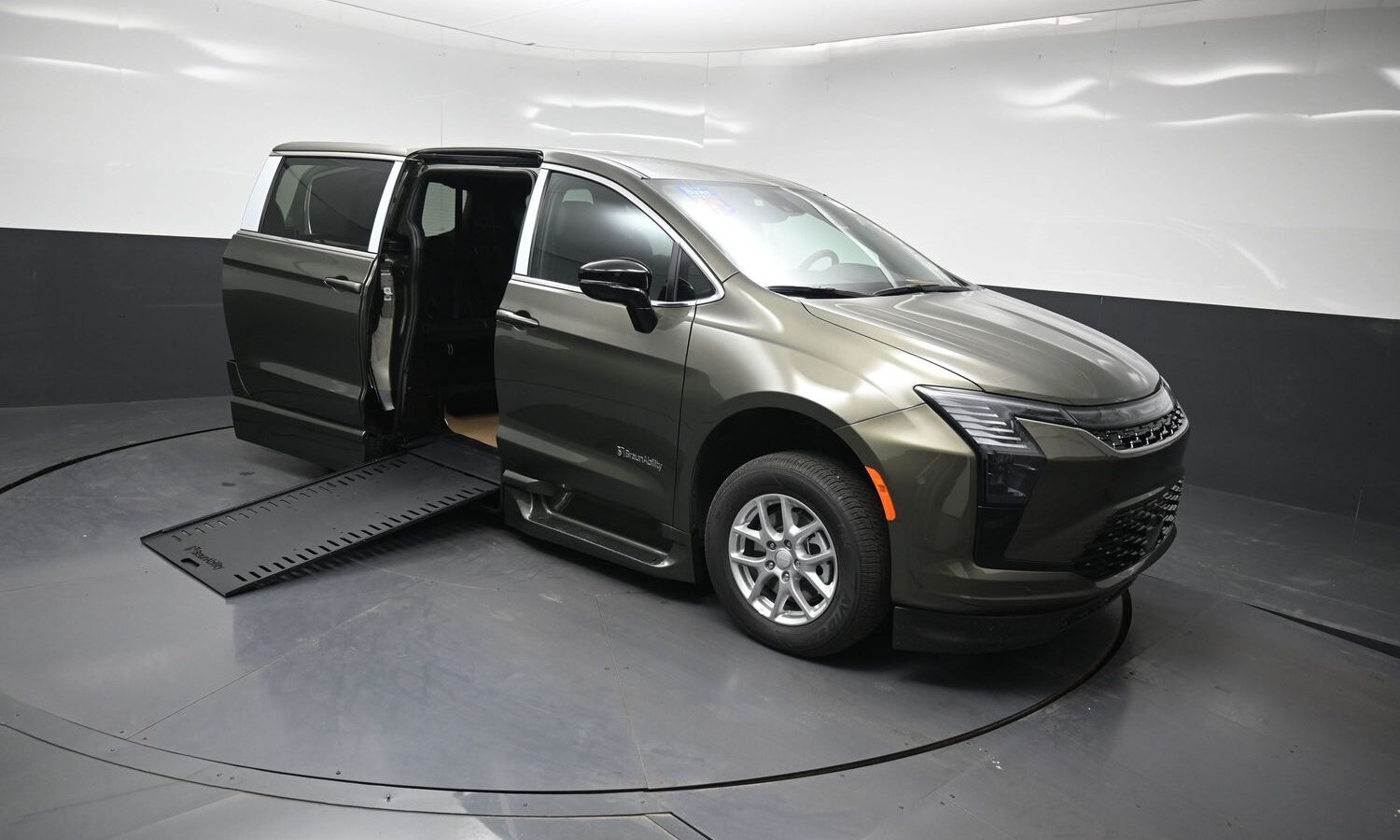 Olive-green minivan with a side-entry ramp extended, door open, displayed on a turntable in a showroom, highlighting accessibility features.