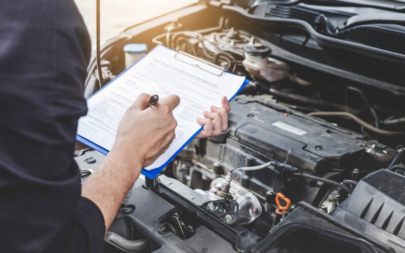 Automobile mechanic repairman checking a car engine with inspecting writing on the clipboard checklist for car service and maintenance.