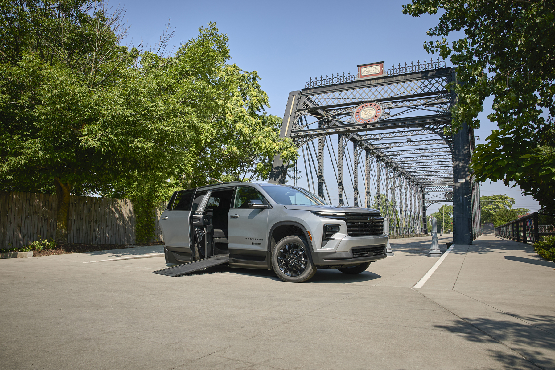 Silver SUV with its side ramp deployed, parked beside a wrought-iron bridge and leafy trees on a sunny day