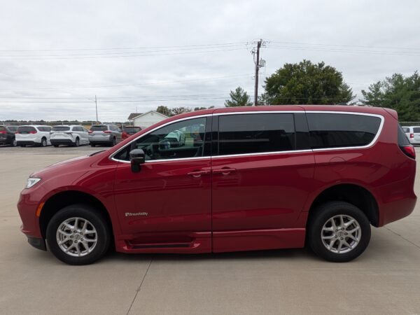 PXL_20260922_160951497 Side view of a red BraunAbility wheelchair-accessible minivan parked in a lot with other vehicles and a cloudy sky in the background.