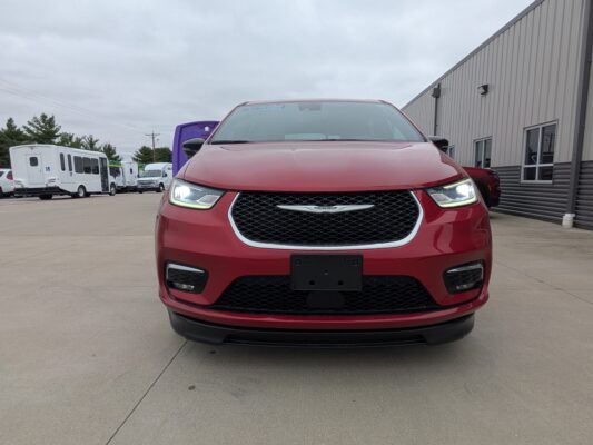 Front view of a red Chrysler SUV facing the camera in a parking lot with a gray industrial building in the background, headlights on
