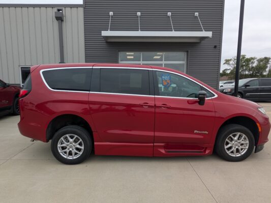 Red minivan shown in profile parked in front of a gray metal building, chrome trim along the windows, BraunAbility badge visible on the rear door.
