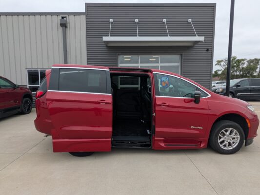 Red BraunAbility wheelchair-accessible van with the side door open in a parking lot in front of a gray industrial building.