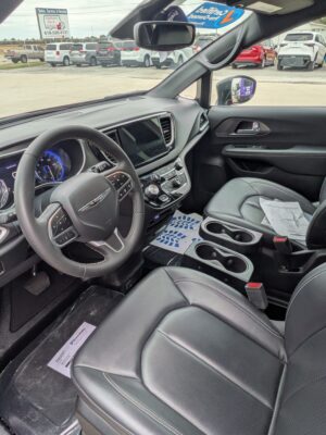Interior of a black SUV: steering wheel, large touchscreen, center console with knobs, and leather seats with stitching; papers on the front passenger seat, dealership lot visible through the windshield
