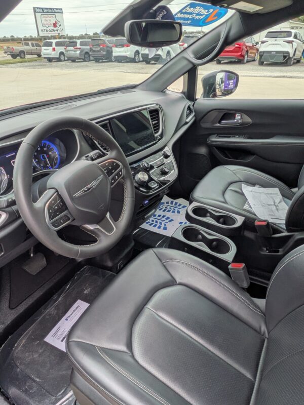 PXL_20260922_161224877 Interior of a black SUV: steering wheel, large touchscreen, center console with knobs, and leather seats with stitching; papers on the front passenger seat, dealership lot visible through the windshield