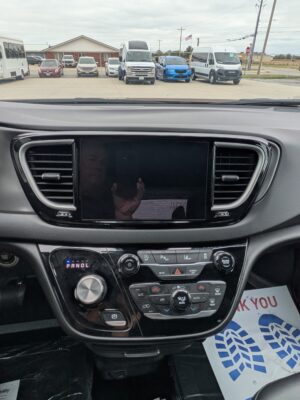 View from the driver's seat of a modern car: center touchscreen, climate controls, gear selector, with a row of vans and cars parked outside through the windshield.