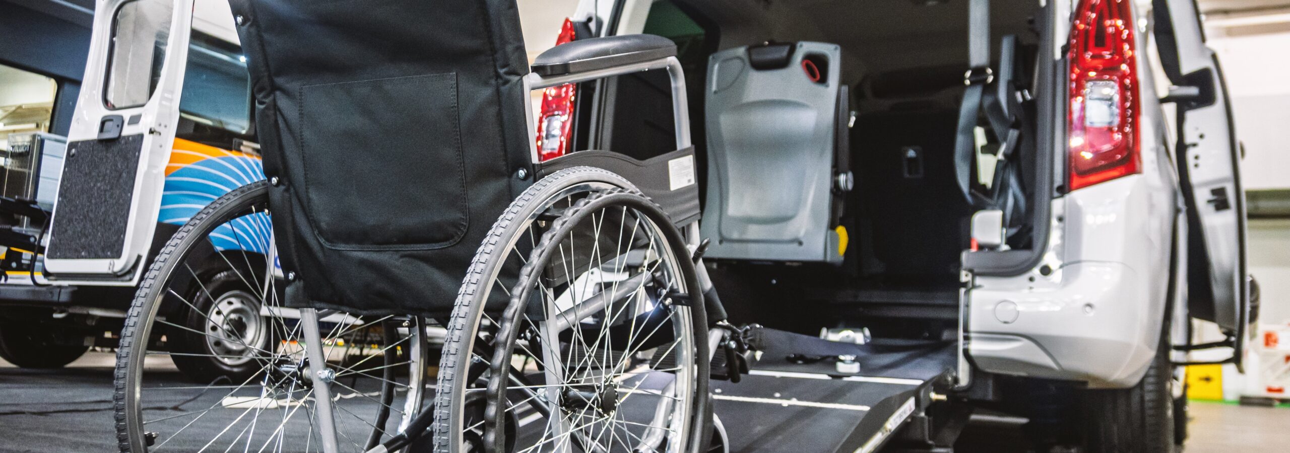 A close-up view of a modern, white accessibility van with its rear door open and a deployable ramp extended, ready for use. A sturdy black wheelchair is positioned in front of the ramp, emphasizing ease of loading and unloading. The interior of the van reveals specialized seating and safety restraints designed for secure transport of passengers with mobility challenges. This image effectively illustrates contemporary solutions for inclusive transportation, catering to individuals with disabilities. It's suitable for various themes including healthcare, disability support, travel accessibility, and automotive adaptations including kneeling suspension.