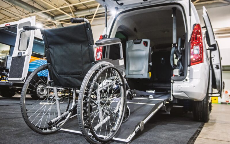 A close-up view of a modern, white accessibility van with its rear door open and a deployable ramp extended, ready for use. A sturdy black wheelchair is positioned in front of the ramp, emphasizing ease of loading and unloading. The interior of the van reveals specialized seating and safety restraints designed for secure transport of passengers with mobility challenges. This image effectively illustrates contemporary solutions for inclusive transportation, catering to individuals with disabilities. It's suitable for various themes including healthcare, disability support, travel accessibility, and automotive adaptations including kneeling suspension.